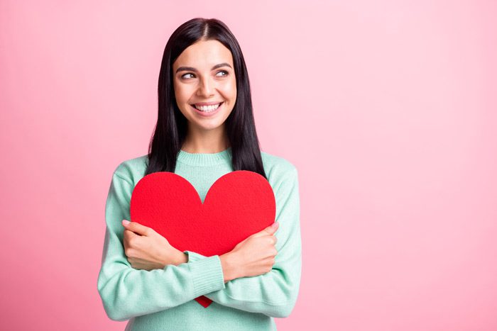 pretty young woman holding a large red heart, and smiling against a pink background - self-love