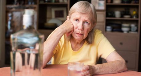 senior woman sitting at a table at home drinking liquor and looking sad - loneliness