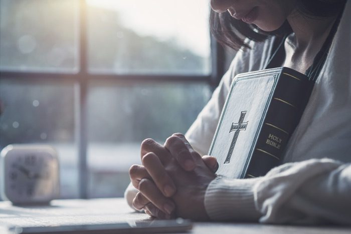 cropped shot of young woman praying with a Holy Bible leaned against her chest - faith-based recovery